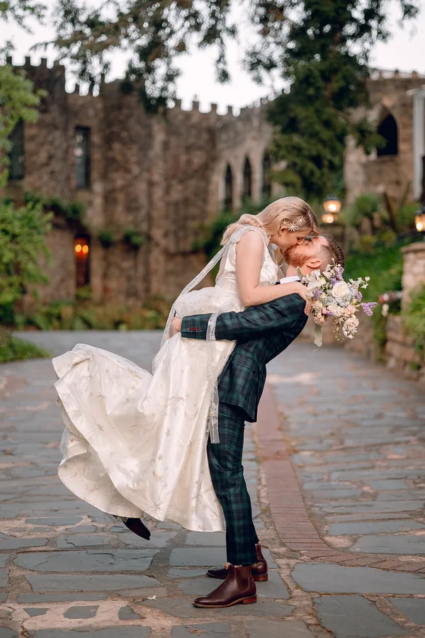 Hitch Perfect — the groom lifting the bride outside a sandstone castle