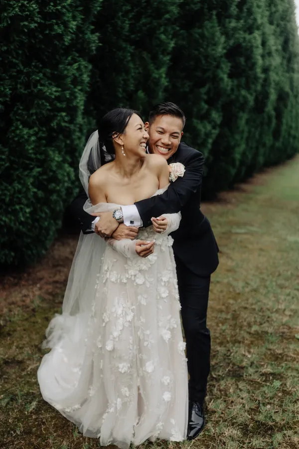 Fable Weddings: Groom hugging his bride from behind in a garden avenue