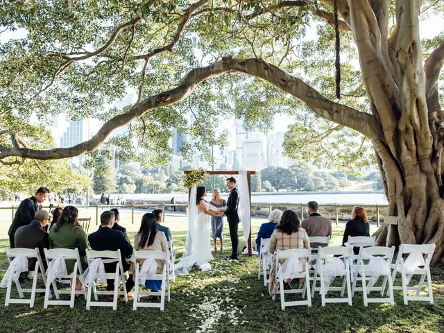 Lovesick Weddings: Ceremony under a fig tree by Sydney Harbour with white padded chairs and a timber arch