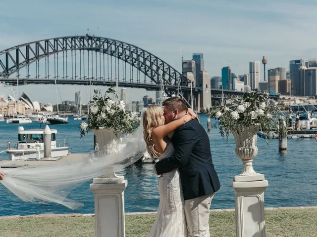 Lovesick Weddings: Newlyweds kiss between floral urns on plinths with the Harbour Bridge behind