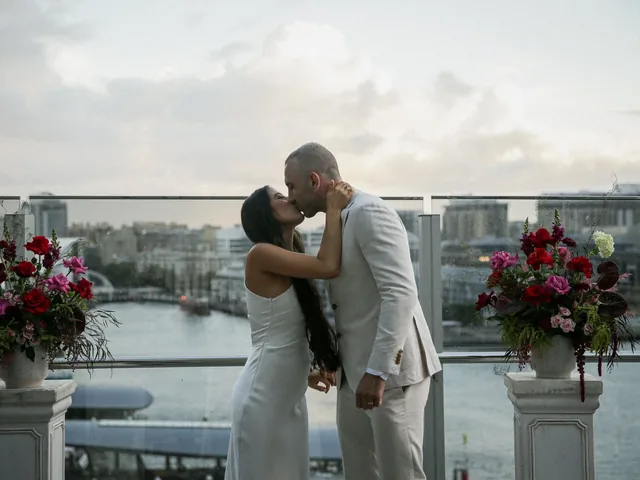 Lovesick Weddings: Couple kiss at a waterfront ceremony framed by red floral urns